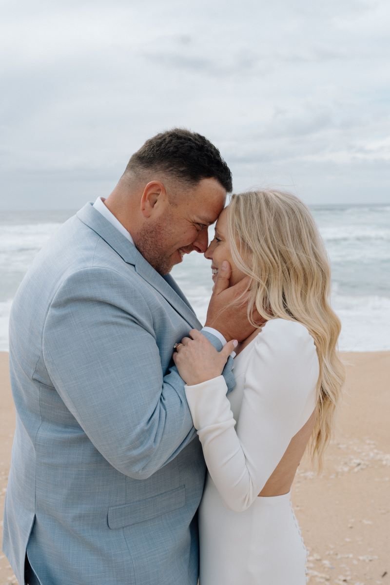 A man and woman on the beach next the ocean they have their heads pressed together and are smiling the woman is wearing a white wedding dress the man is wearing a blue suit and holding the woman's face in his hands 