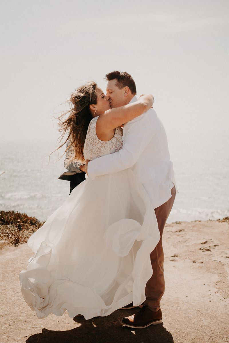 A man and woman kissing on a cliff by the ocean the woman is wearing a white wedding dress and hugging the man around his shoulder the man is wearing a white suit jacket and tan pants and hugging her around the waist 