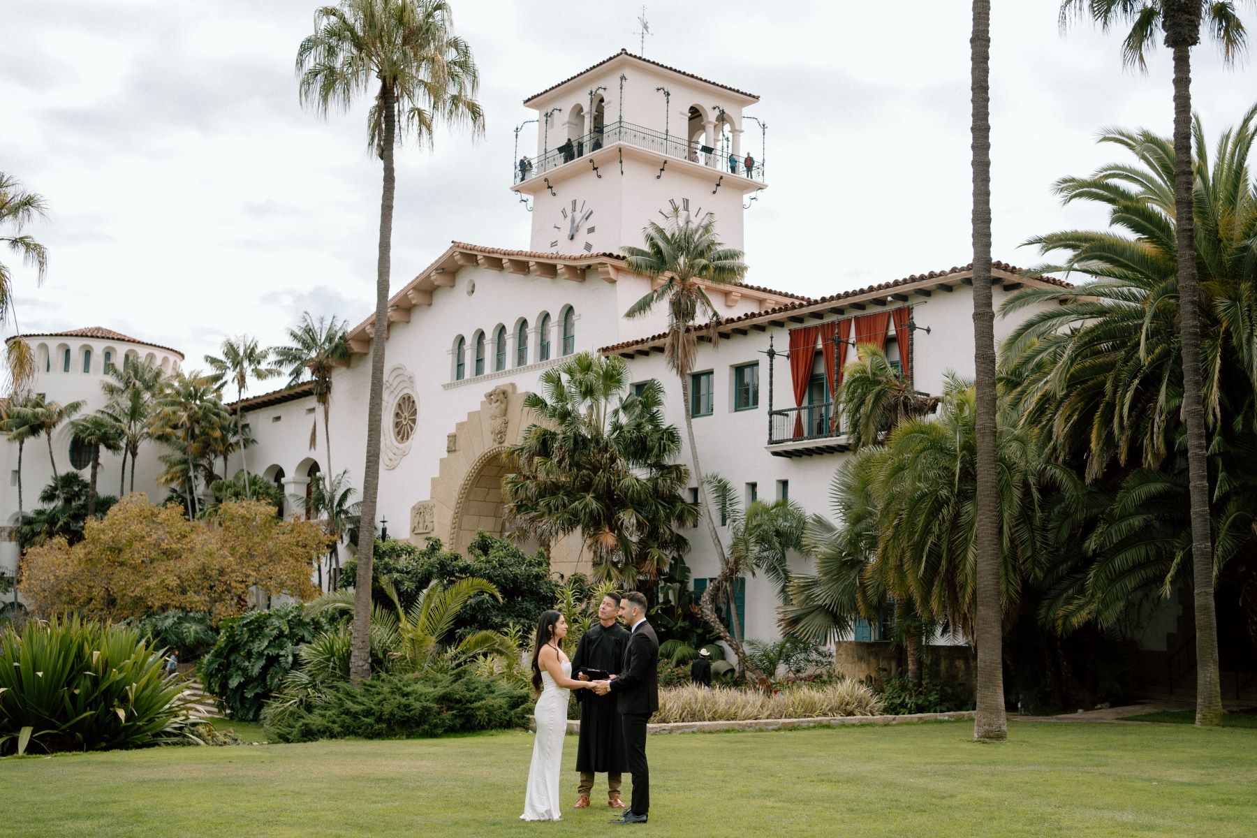 A couple at their elopement ceremony with their officiant holding hands and looking at each other the woman is wearing a white wedding dress the man is wearing a suit behind them are plants, palm trees, and a white spanish colonial style hacienda 