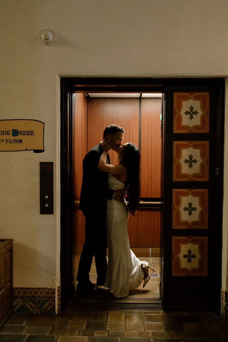 A couple inside of an elevator kissing the woman is wearing a white wedding dress and kicking up one heel the man is wearing a black suit 