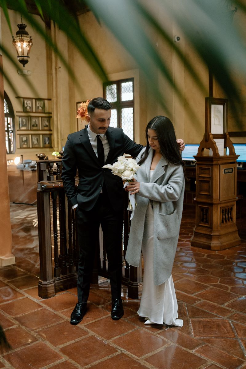 A couple in a courthouse the woman is wearing a gray jacket and white wedding dress and holding a bouquet of flowers and her partner is wearing a black suit and is putting his arm around her