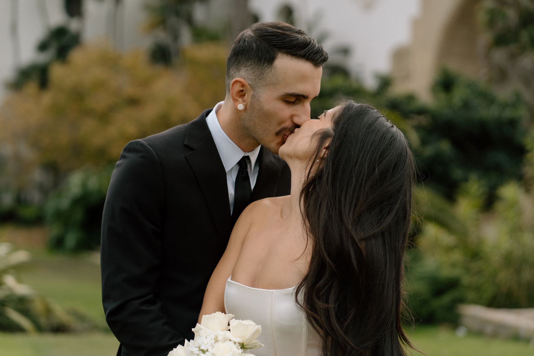 A couple kissing each other the woman is wearing a white wedidng dress and holding a bouquet of flowers the man is wearing a black suit 