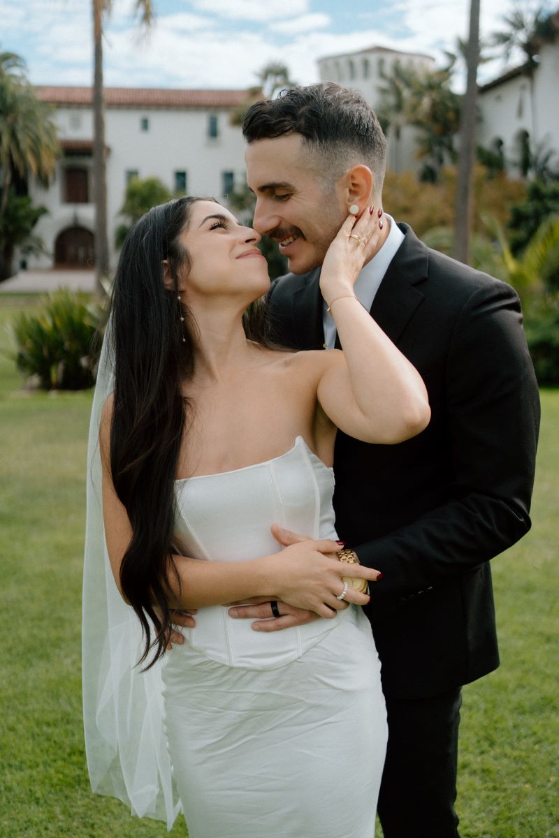 A couple looking into each other's eyes the woman is looking back at her partner and touching his face with one arm with the other she is clasping his hand the woman is wearing a white wedding dress and veil and the man is wearing a black suit 