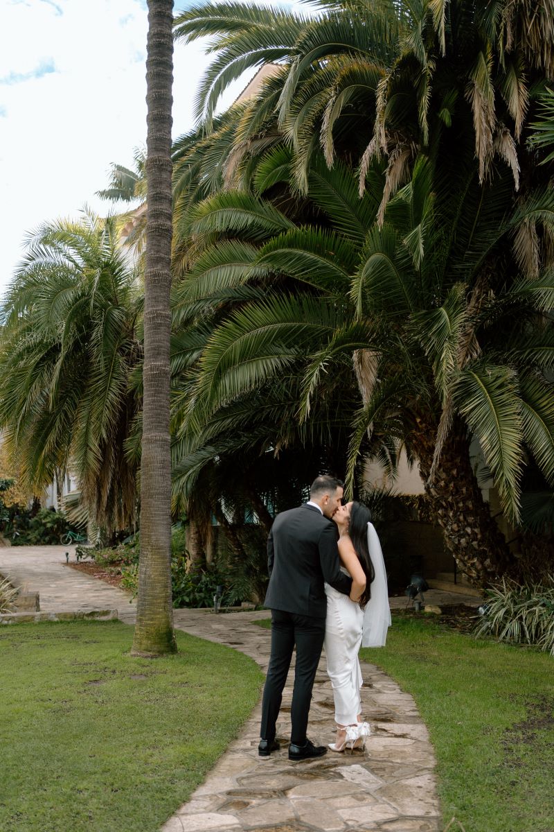 A couple kissing each other underneath palm trees the man has his arm around her waist the woman is wearing a white wedding dress and veil and the man is wearing a black suit