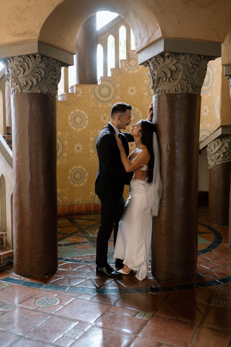 A couple inside of a courthouse looking into each other eyes the woman is leaning back against a pillar and has her hand on her partner's chest 