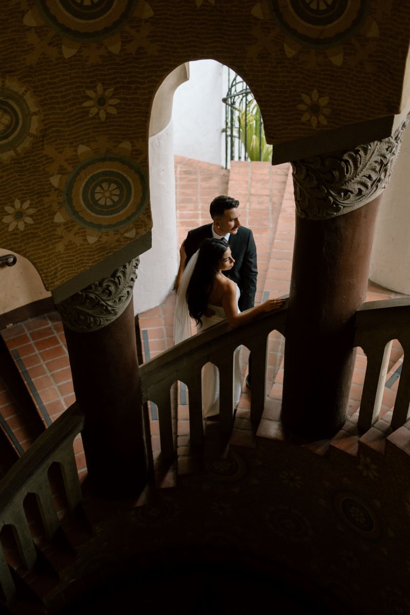 A couple inside of a courthouse next to a spiral staircase the man is wearing a black suit the woman is wearing a white wedding dress and veil 