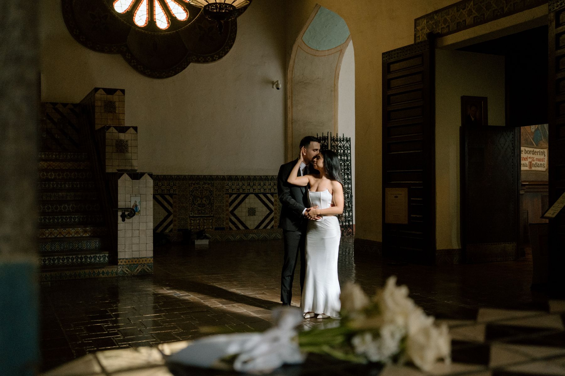 A couple inside of a room inside of the santa barbara courthouse the man is hugging his partner from behind and kissing the side of her head the woman is wearing a white dress and touching her partner's head with one hand with the other she is holding her partner's hand 