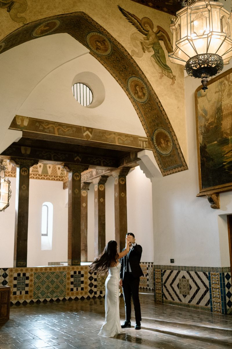 a couple dancing in a large room inside of the santa barbara courthouse the woman is wearing a white dress the man is wearing a suit and spinning her 