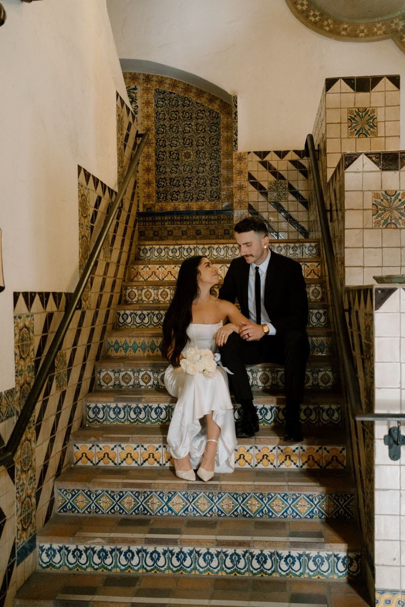 a couple sitting together on a staricase the woman is wearing a white dress and holding a bouquet of folowers the man is touching her elbow and wearing a black suit and is sitting behind her