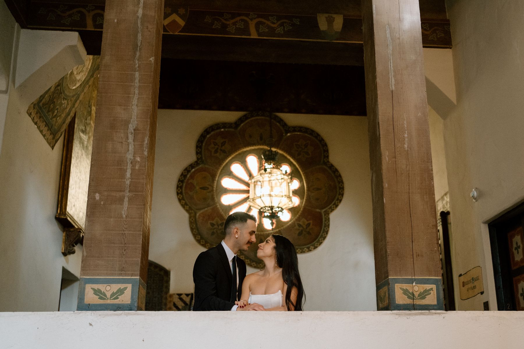 A couple at a courthouse after their wedding ceremony standing next to a railing looking at each other and smiling they are surrounded by wood columns and behind them is a latern hanging from the ceiling 