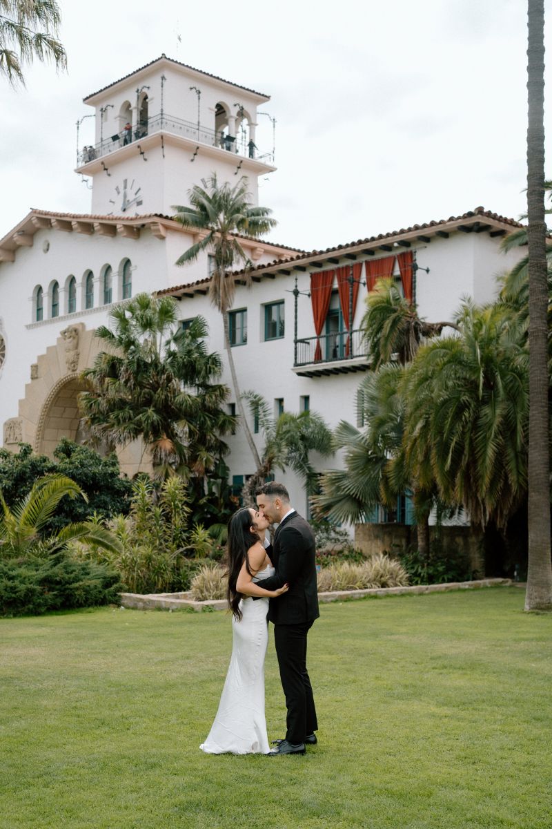 A couple having their wedding ceremony in front of the santa barbara courthouse they are both kissing one another the woman is wearing a white wedding dress the man is wearing a black suit 