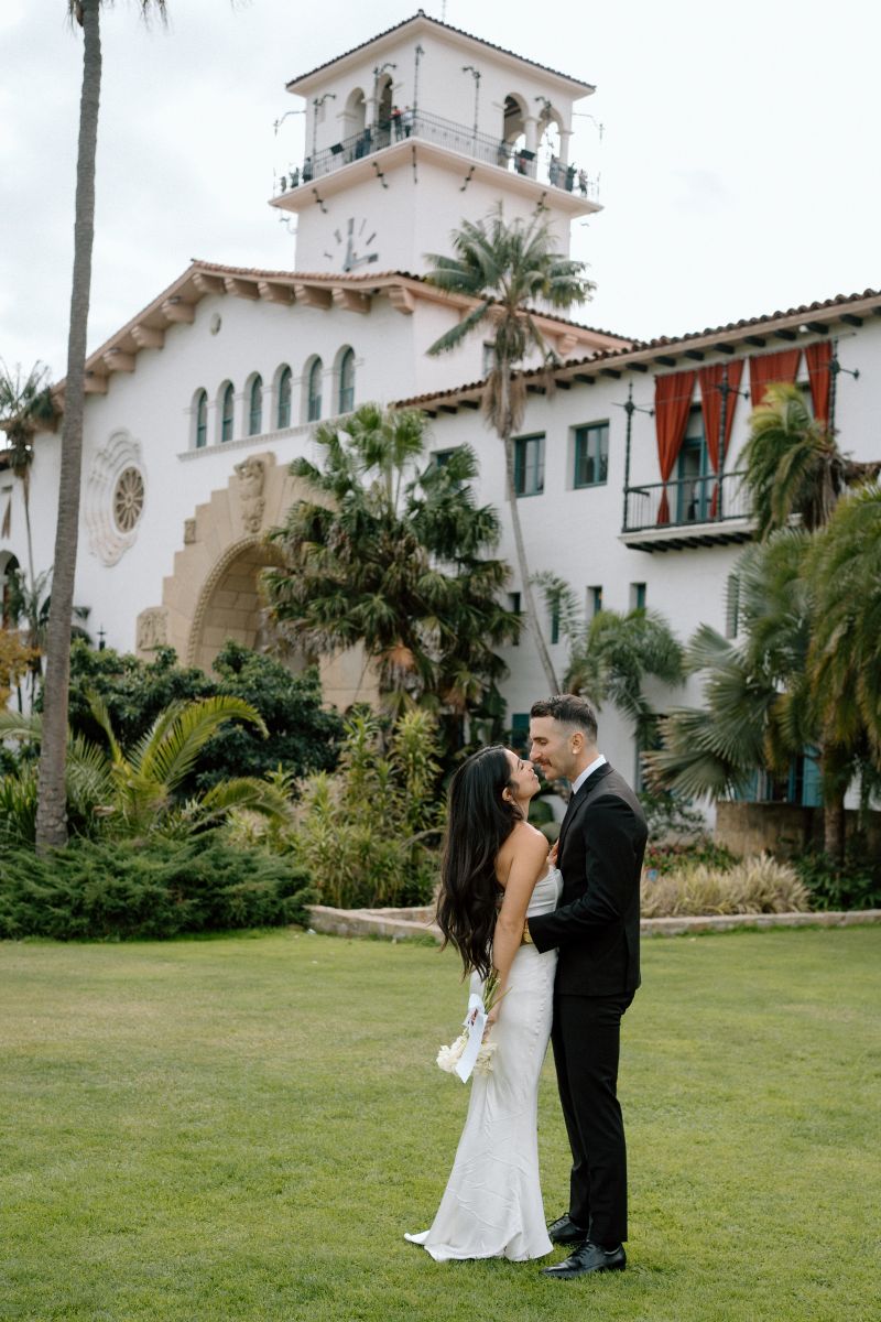 A couple having their wedding ceremony in front of the santa barbara courthouse one another the woman is wearing a white wedding dress the man is wearing a black suit the woman is also holding a bouquet of flowers and the man is grabbing her around the waist they are looking at each other and smiling