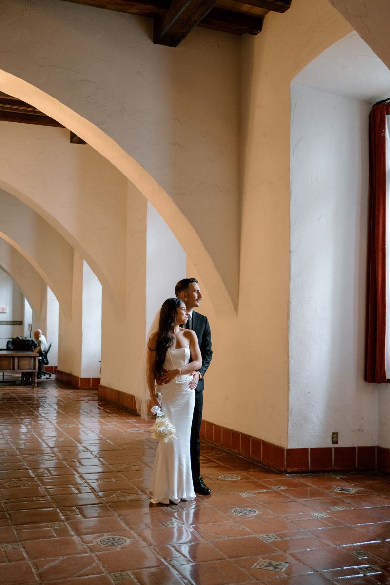 A couple standing together inside of a courthouse the man has his arms around the woman's waist and is holding her from behind the woman is wearing a white wedding dress and holding a bouquet of white flowers 