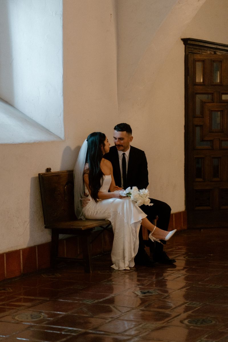 a couple sitting together on a wooden bench inside of a courthouse the woman is wearing a white veil and white dress and holding a bouquet of flowers the man is wearing a black suit the couple is looking at each other