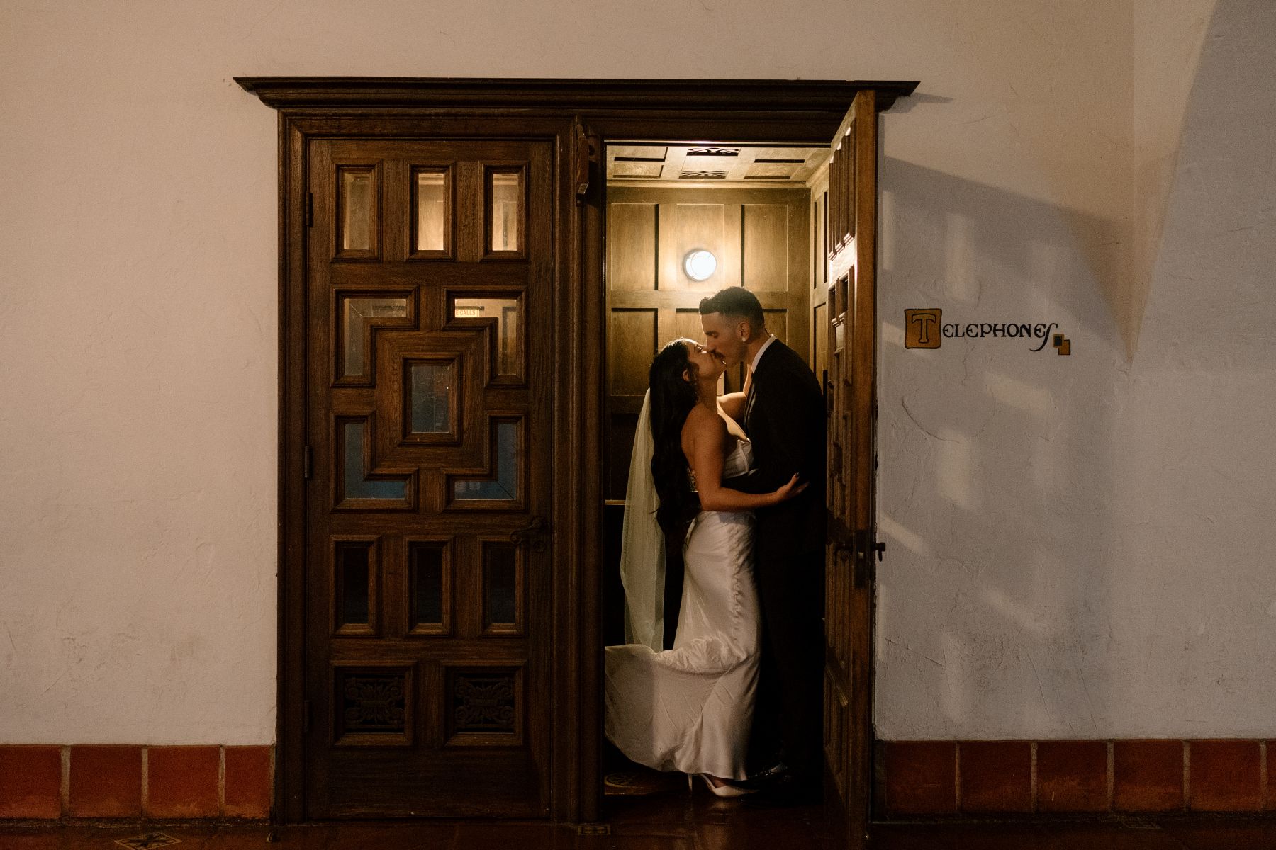 A couple insideo fo a courthouse standing in the archway on an old telephone booth the man is kissing the woman who is wearing a white dress and veil the man is wearing a black suit