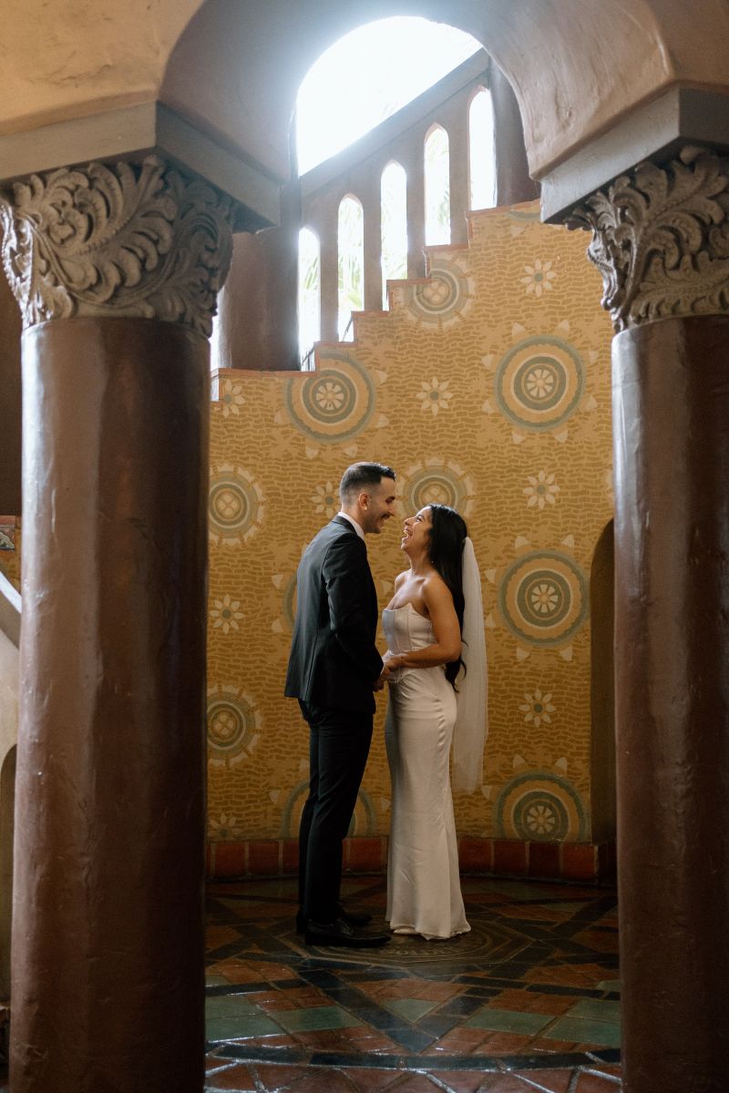 A couplie inside of the santa barbara courthouse the woman is wearing a white dress and white veil and holding her partner's hand and laughing the man is wearing a black suit and laughing with her 