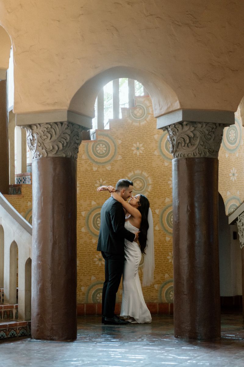 A couple inside of the santa barbara courthouse next to two large columns and a large staircase the woman is holding her partner around the shoulders and the man is hugging the woman around her waist 